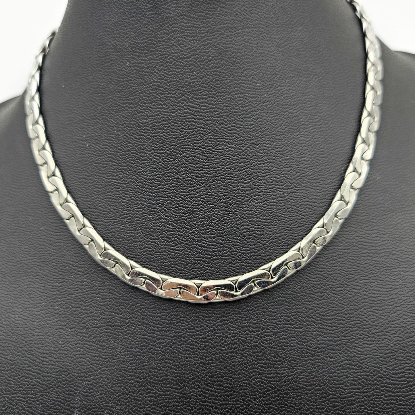 A silver-toned metal chain necklace displayed on a black textured jewelry bust against a white background.