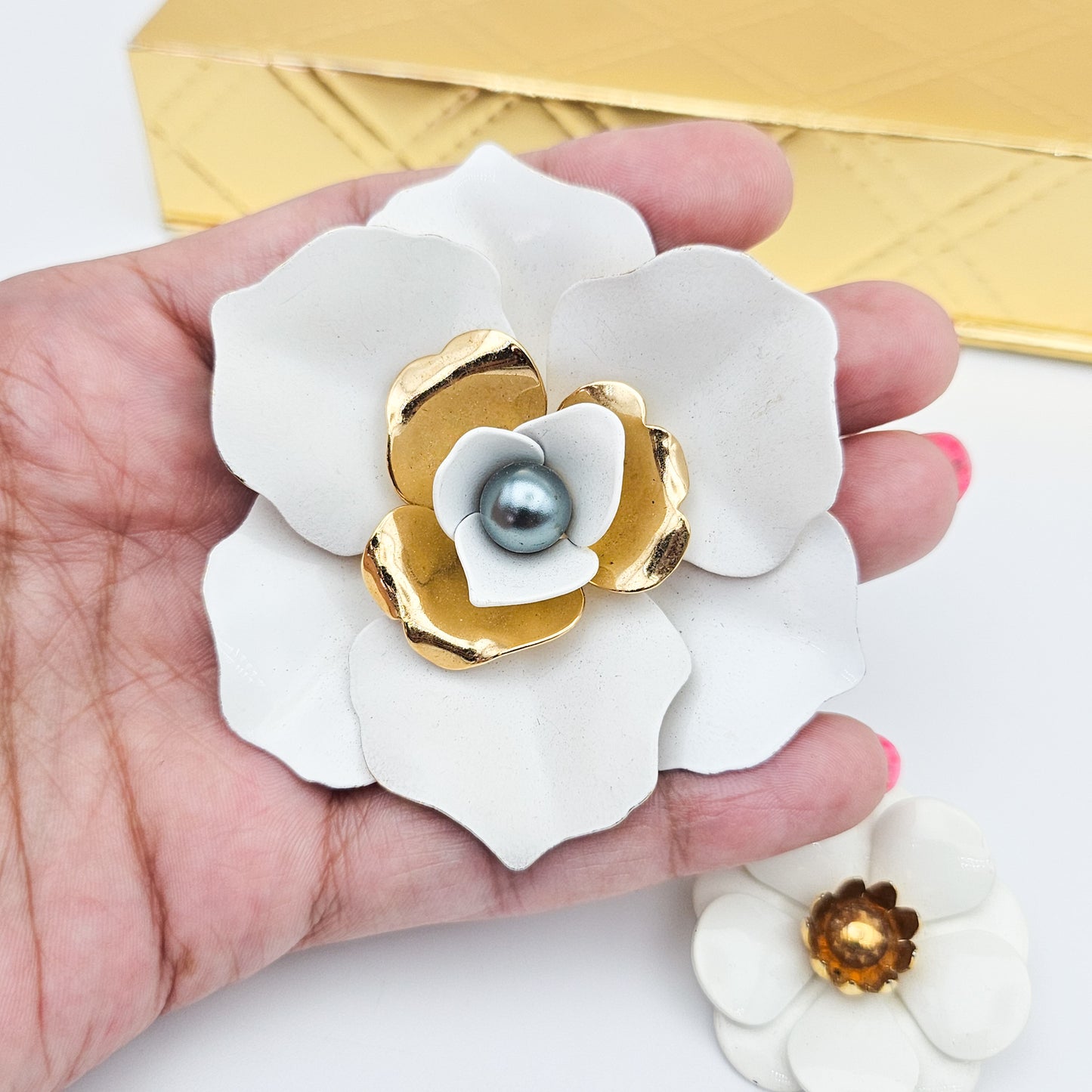 A hand holding a Signed Sarah Coventry white enamel flower brooch with gold-tone inner petals. A smaller coordinating flower pin sits on the surface nearby.
